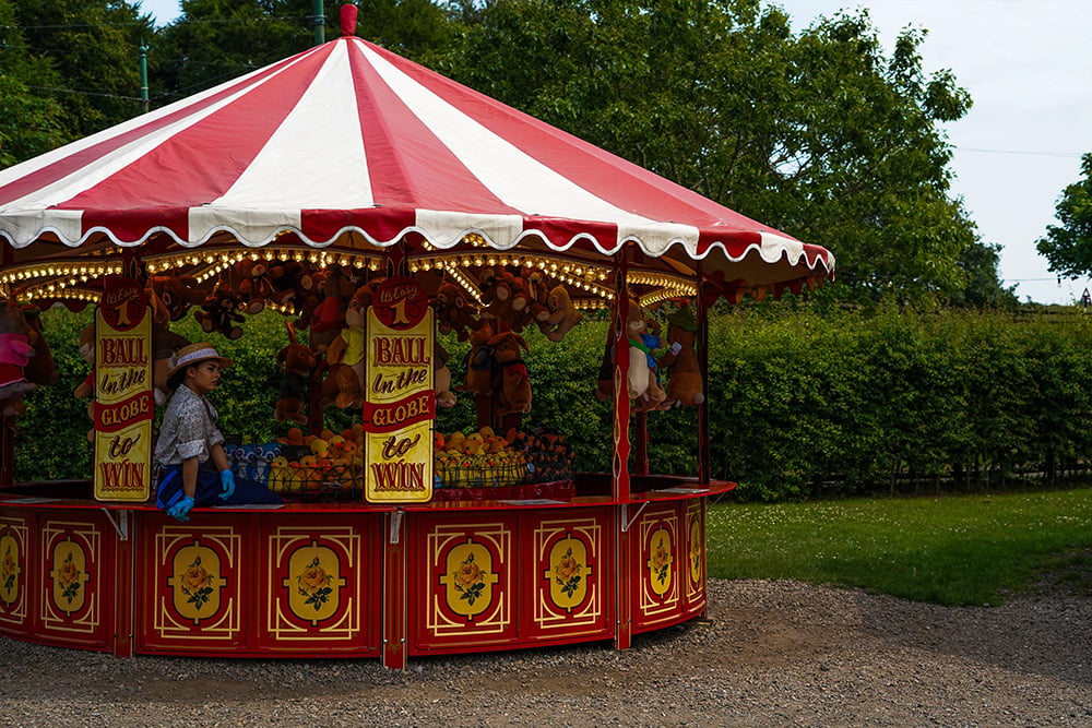 Colorful vintage carnival game stall with a red-and-white striped canopy, ornate red panels, plush prizes, and “Ball in the Hole” signs.